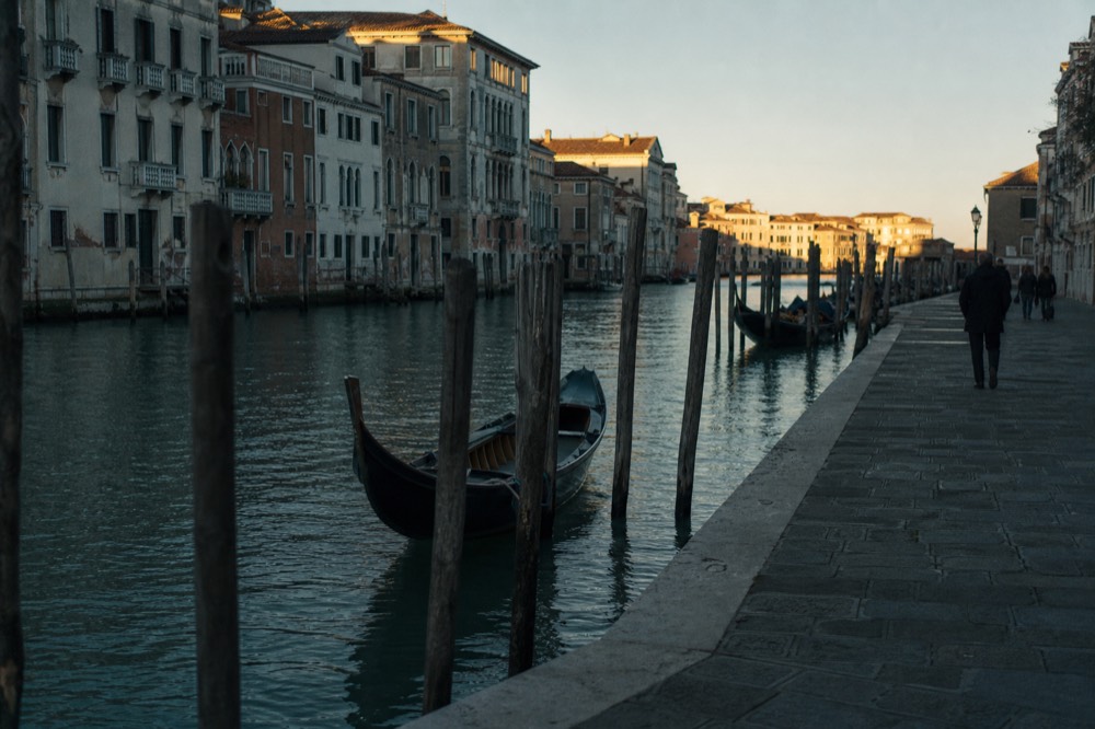 Low winter sun striking the upper windows of Venetian palaces above still canal water, long shadows across a stone quay