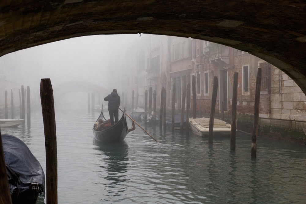 Winter fog over a Venetian canal with mooring poles fading into grey and a bridge outline barely visible ahead