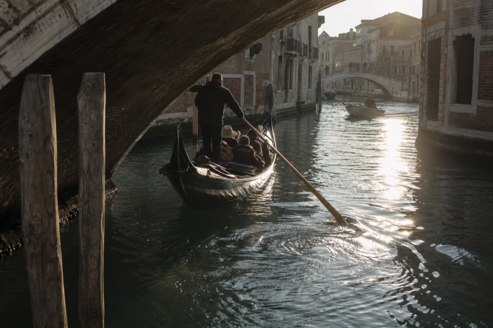 A gondola turning tightly at a canal junction beneath a low stone bridge, the oar mid-stroke and rings of water spreading behind it