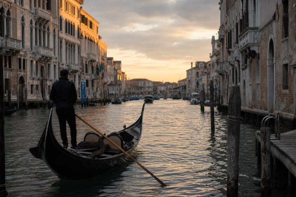 A gondolier standing idle at the stern with his oar laid across the boat, waiting beside an empty mooring on a sunlit canal