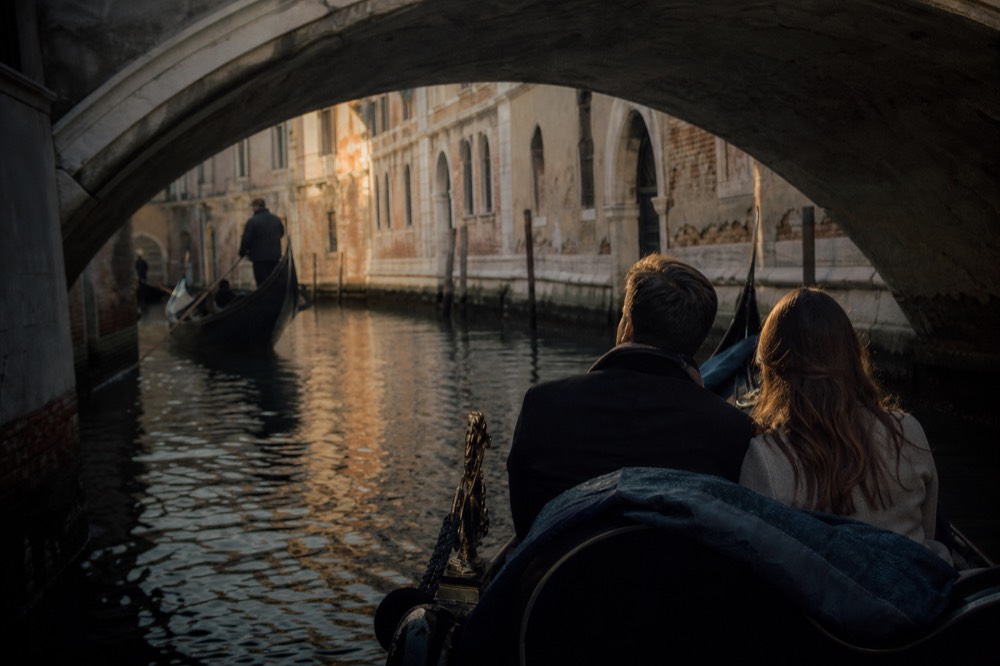 Two passengers seated close together on a private gondola gliding under a low stone bridge in a narrow shaded canal