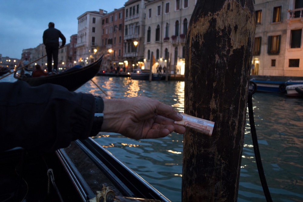 A weathered hand passing a folded euro note across a boat gunwale toward a mooring post, water blurred behind