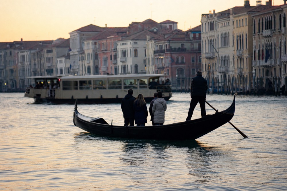 Passengers standing upright in a plain working gondola crossing a wide canal, a crowded public waterbus passing behind them