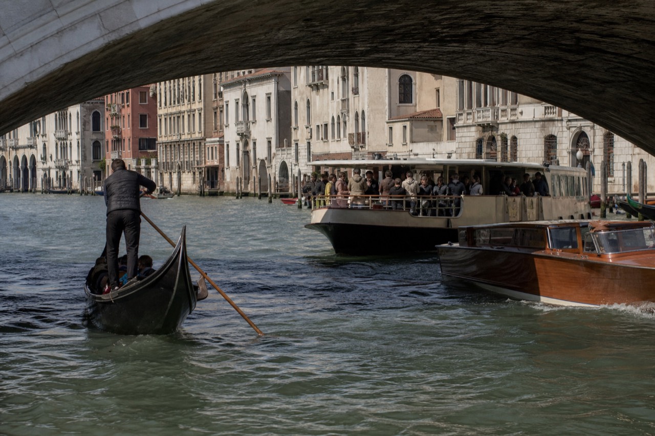 Three kinds of Venetian boat sharing the same stretch of canal, a rowed black hull, a crowded public waterbus and a low varnished launch