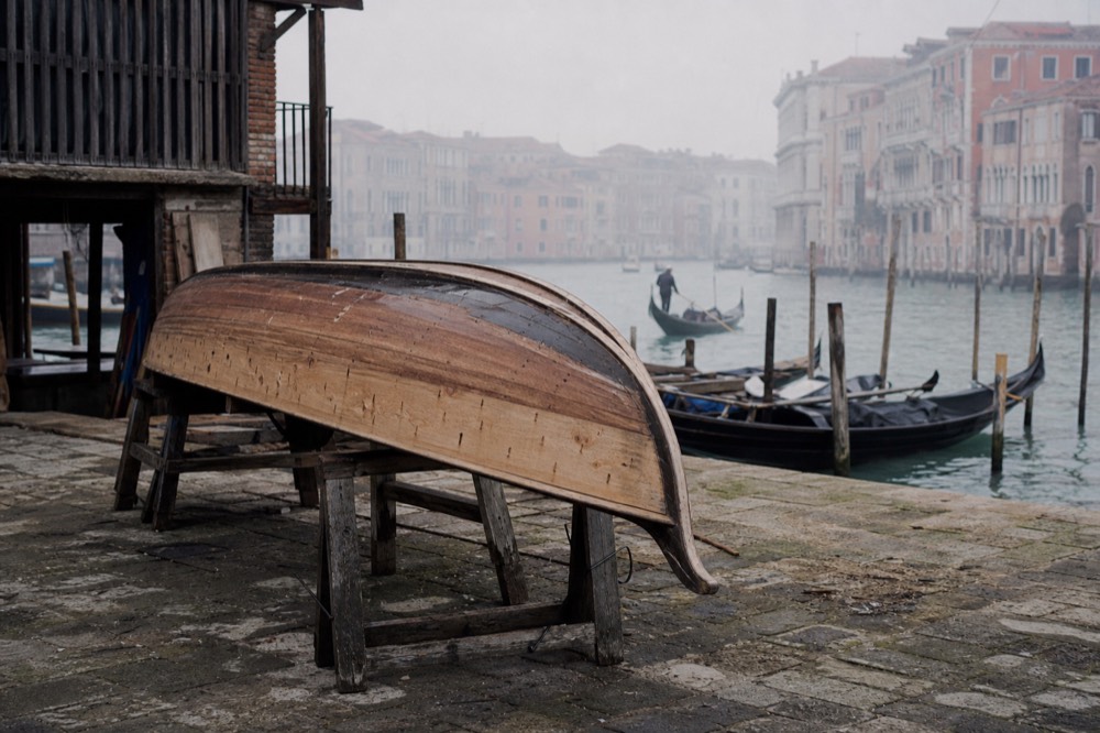 An upturned gondola hull resting on trestles in an open boatyard, bare timber planking visible along one flank