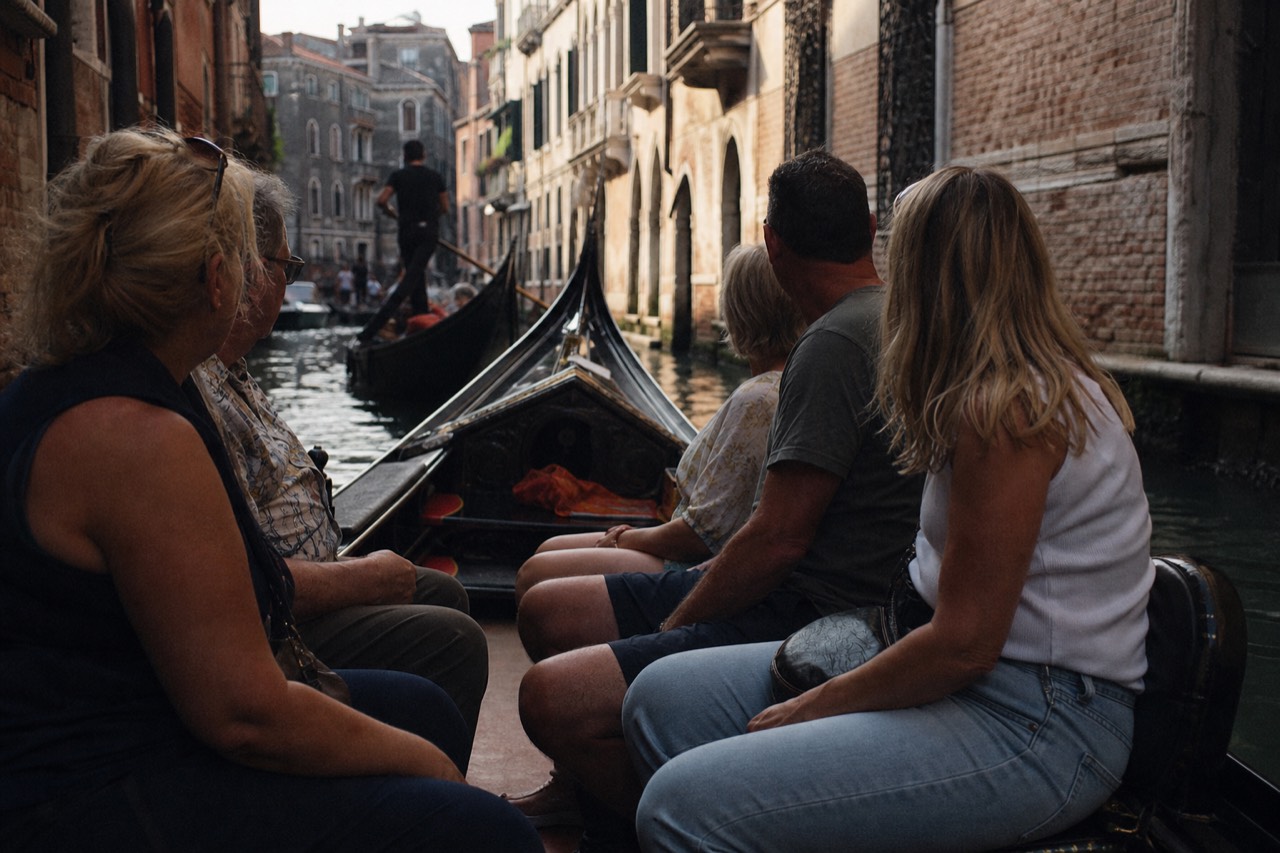 Five passengers seated along the black side benches of a gondola gliding through a narrow Venetian canal in flat afternoon light