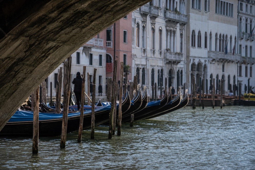 A row of moored gondolas rocking side by side in front of pastel palazzo facades on a wide Venetian waterway at midday