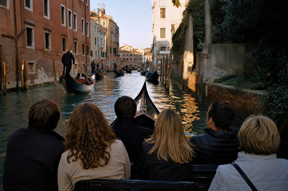 Six seated passengers viewed from behind on a shared gondola bench as it turns from a busy waterway into a shaded back canal