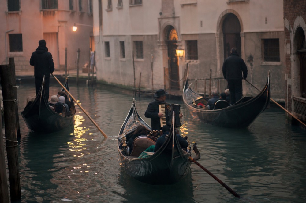 A small cluster of gondolas drifting close together at dusk while an accordion player performs from the middle boat, lamplight on the water