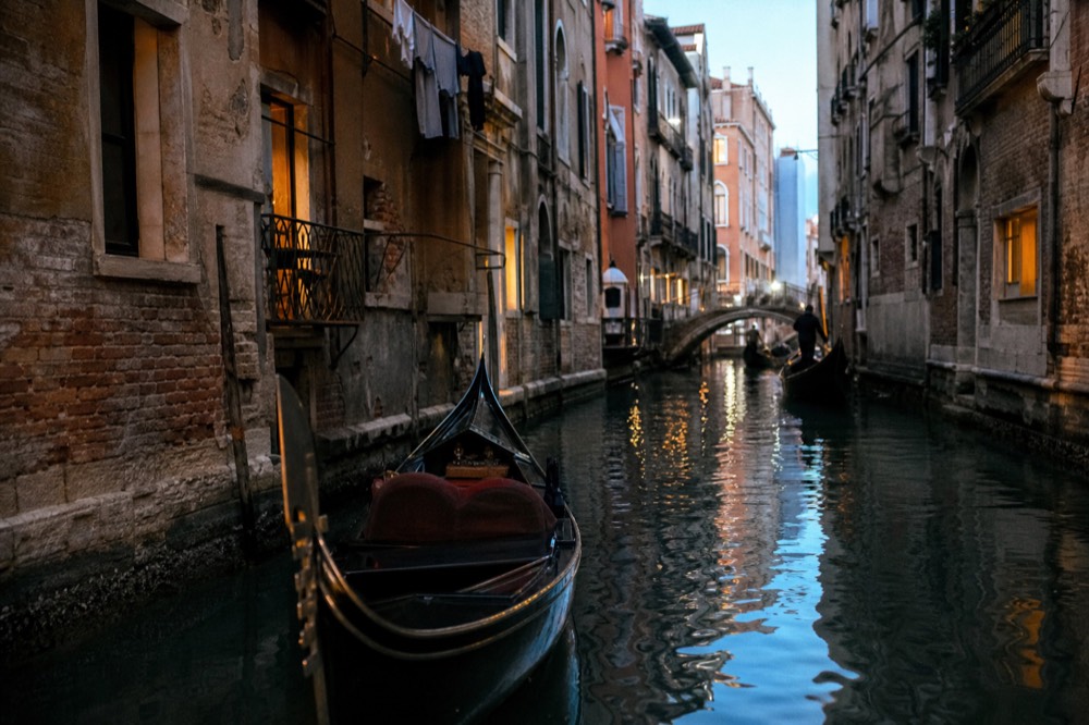 An empty gondola gliding through a narrow back canal in soft late light, laundry lines and iron balconies overhead on both sides