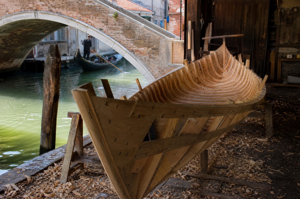 A half built wooden gondola hull resting on trestles inside an open boatyard shed, curled wood shavings scattered on the floor