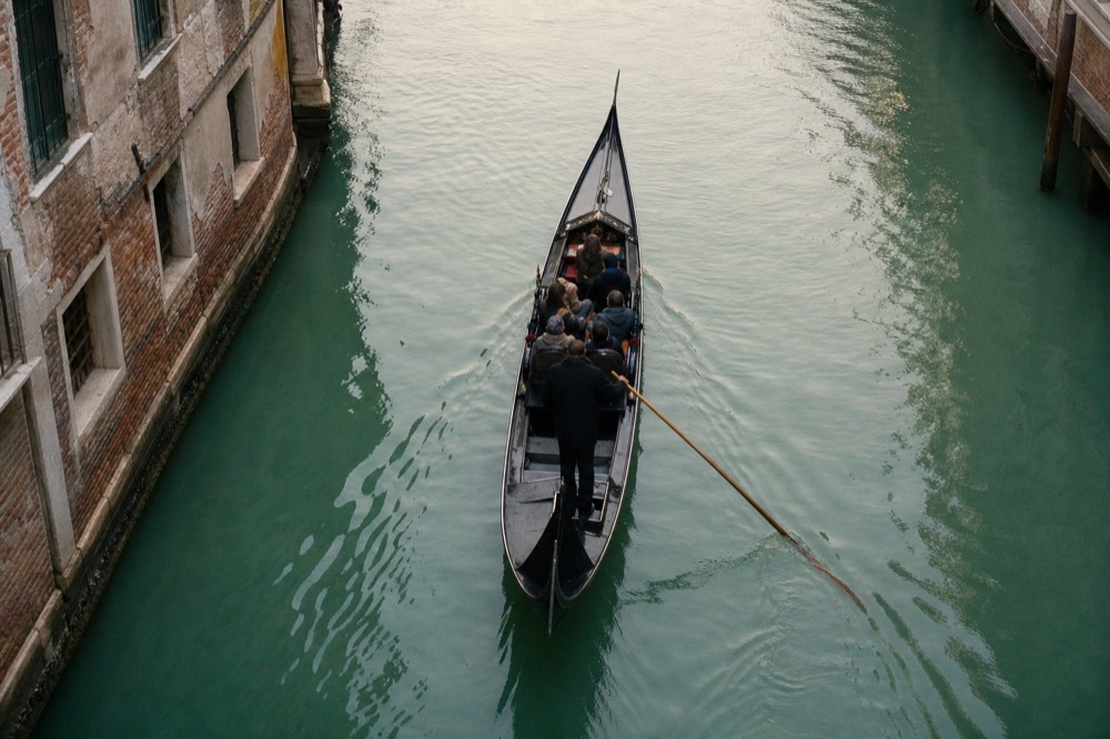 Five passengers seated along the bench of a single gondola on a wide green waterway, photographed from a bridge directly above at midday