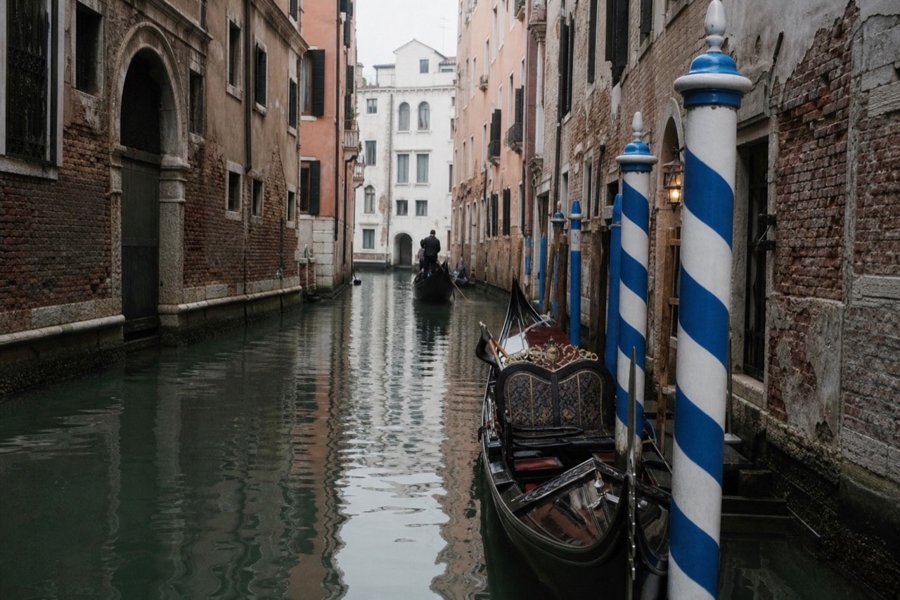 A black gondola moored to blue and white striped poles on a narrow side canal at first light, damp brick walls rising on both sides
