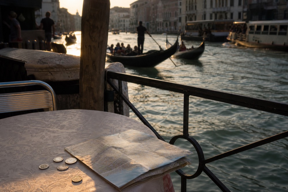 Coins and a folded paper map on a cafe table beside a canal railing, boats moving in soft focus beyond