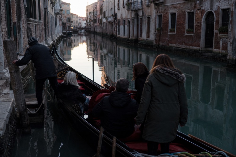 Five people settling onto the cushioned seats of a single gondola while the gondolier unties a rope from a mooring post
