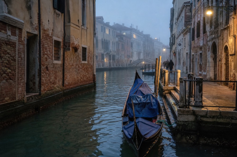 An empty gondola tied alongside a quiet canal wall at dusk with lamp light pooling on the water beside it