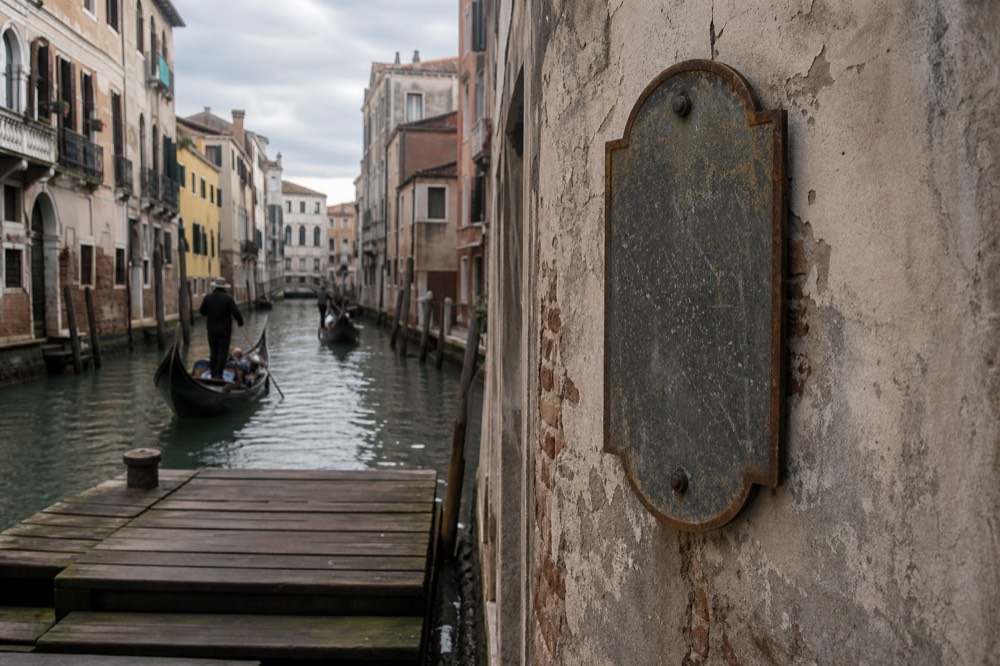 A worn metal price sign bolted to a plastered canal wall beside a wooden landing stage, no readable text, soft overcast light