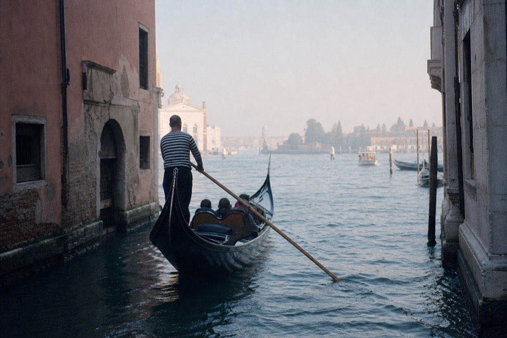 A gondolier in a striped shirt standing at the stern oar as his boat turns out of a side canal into wide open water, seen from behind