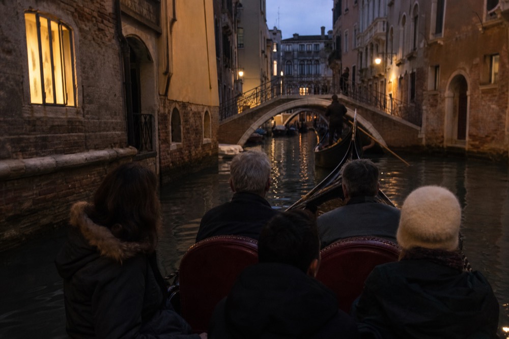 Five passengers seated on the cushioned bench of a gondola from the water level, faces turned away toward a bridge ahead