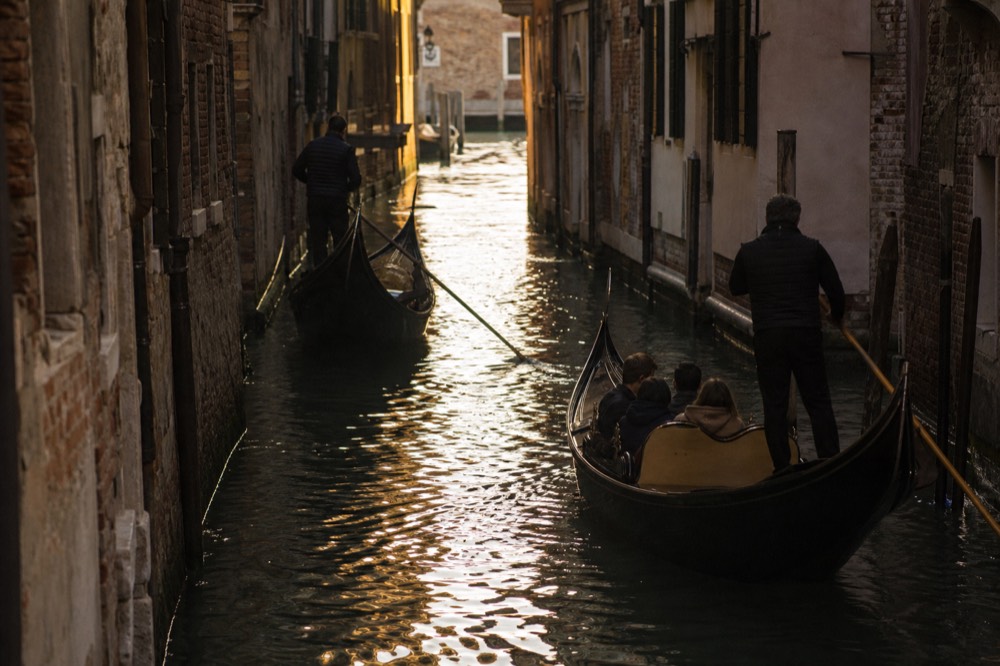 Two gondolas passing in opposite directions in a narrow shaded canal, one carrying a full bench of passengers and the other empty