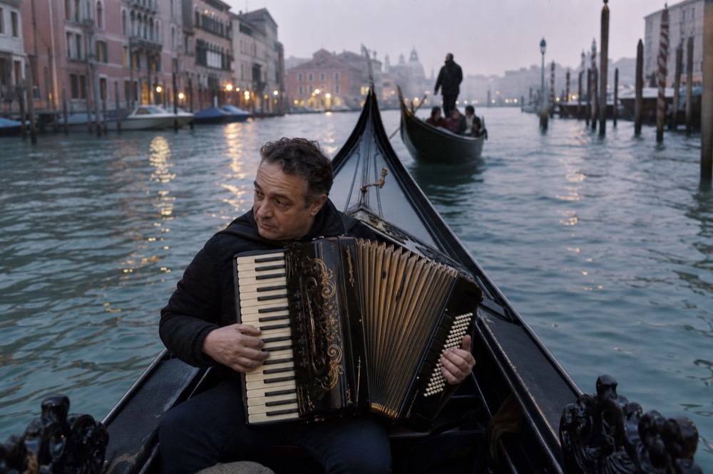 An accordion player seated in the bow of a gondola with the instrument open across his lap, evening light on the water behind him