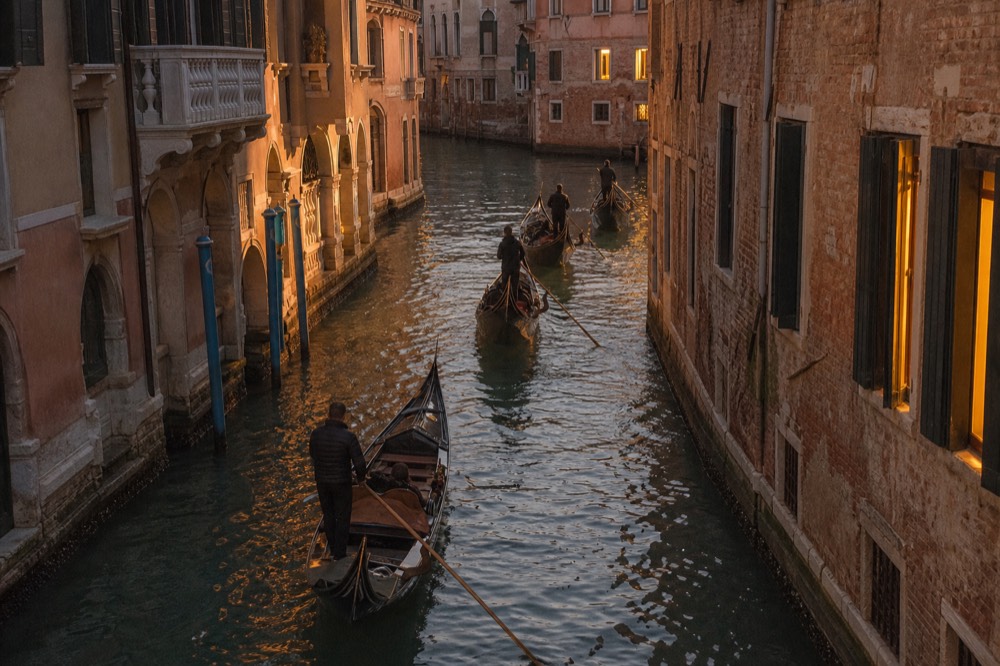Four gondolas photographed from a bridge travelling nose to tail down a canal at twilight, warm window light reflected in the ripples between them