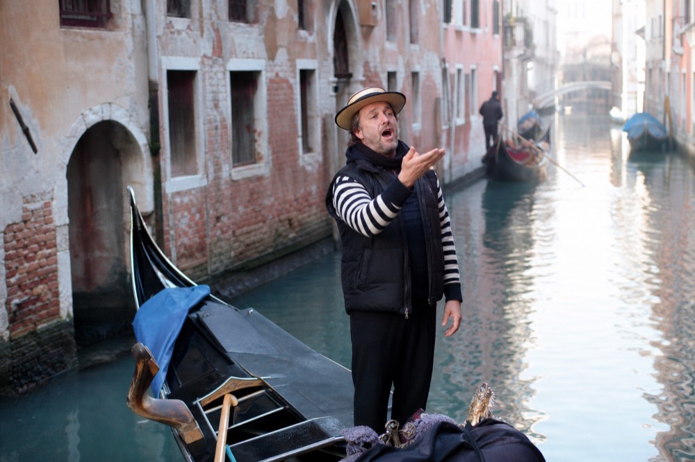 A singer standing braced in a moving gondola mid-phrase, one hand raised, the canal behind him falling into blue evening shadow