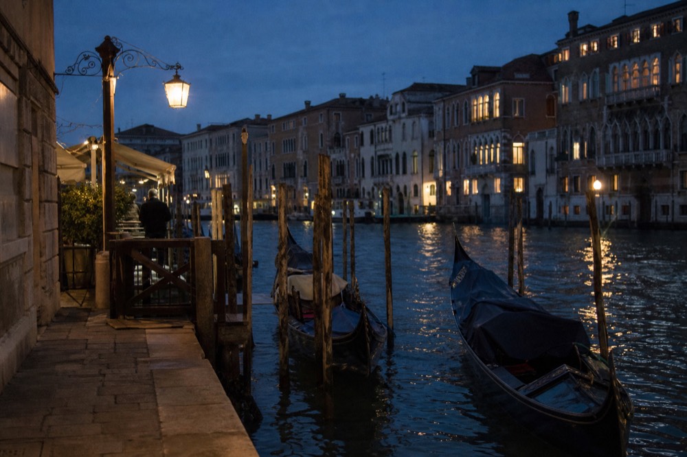 A gondola station on a stone embankment at dusk with mooring posts, a hanging lantern and two moored boats waiting empty