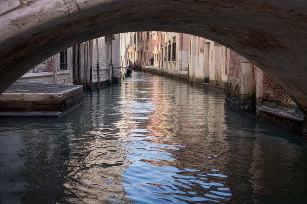 Reflections of lit windows breaking across dark canal water in the wake of a passing boat, no vessels visible in frame
