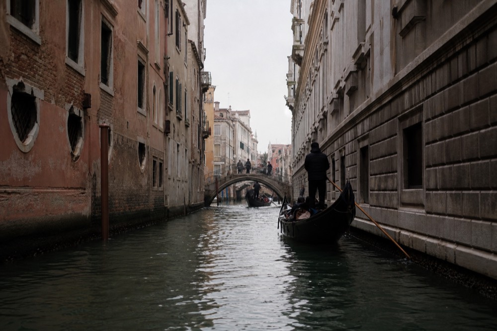 A narrow canal seen at eye level from water height, walls closing in on both sides, a small arched bridge silhouetted ahead against pale sky