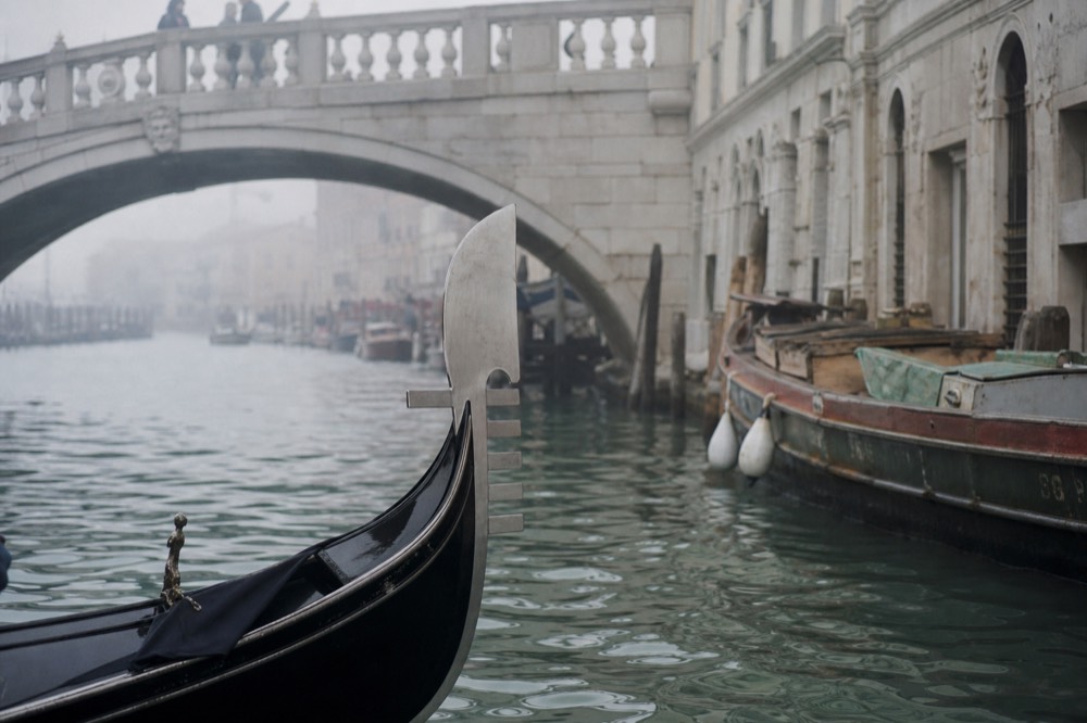 A gondola prow with its steel comb ornament in the foreground, a stone bridge arch and moored working boats beyond in soft early light