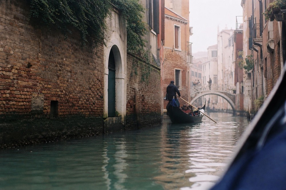 A narrow back canal in Venice with brick walls close on both sides, an arched water gate and a small stone footbridge ahead