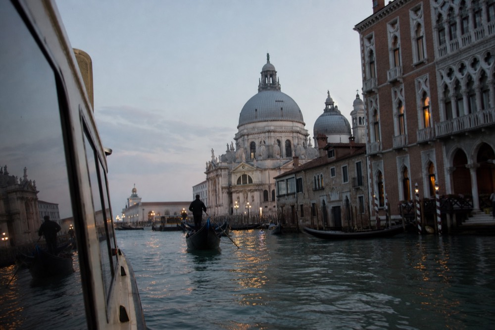 Domed white church rising above the water at the mouth of a Venetian canal, seen low from a passing boat in late afternoon haze