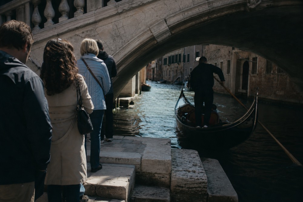 A short queue of travellers standing on a stone landing beside water while a gondolier steadies his boat with one hand on a pole