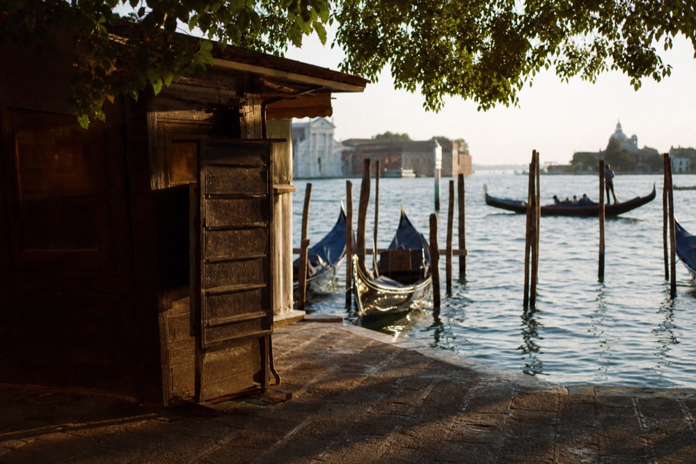 A small wooden departure kiosk beside water with a hand-lettered board of times, seen at an angle in dappled shade