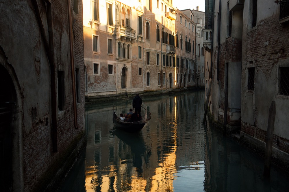 Late golden light striking the upper windows of canal-side buildings while a single gondola glides through shadow below