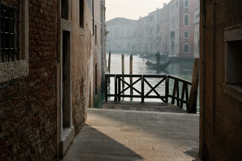 A narrow lane opening onto a small canal with a wooden landing platform and iron railing at the end, morning shadow across the paving