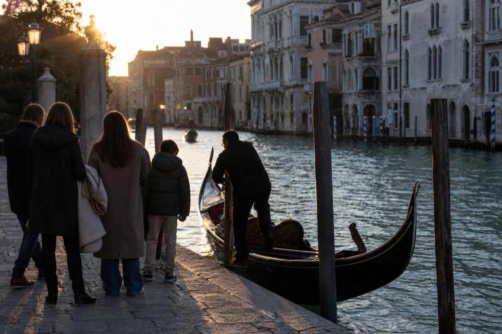 A gondolier stepping onto the stern of his boat while a family waits on the quay with coats over their arms, low winter sun behind