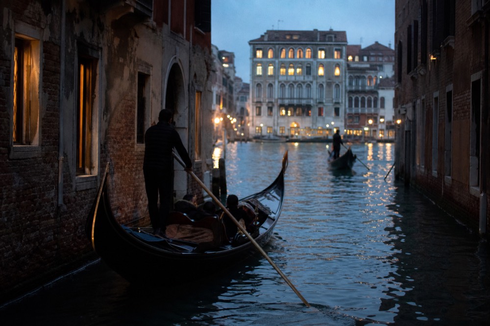 A gondola turning from a shaded side canal into brighter open water, its curved iron prow catching light, weathered shutters above the waterline