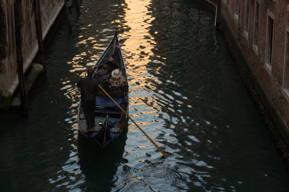 Two people seated alone in a gondola seen from a bridge above, the boat crossing a patch of reflected golden building light on dark water
