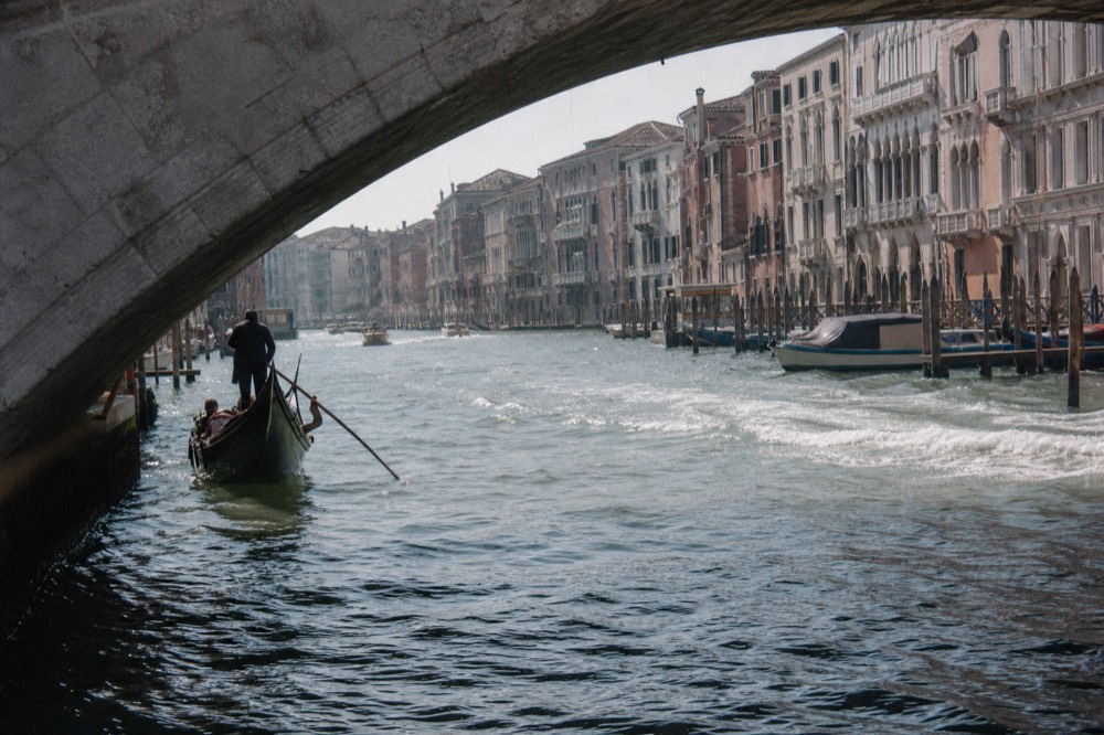 Choppy grey-green water on a broad Venetian waterway with a wake crossing diagonally, palazzo facades receding into haze behind
