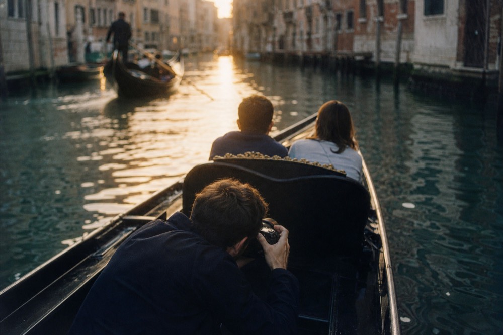 A photographer crouched low in the bow of a small boat aiming a camera back towards two seated passengers, canal walls blurred behind