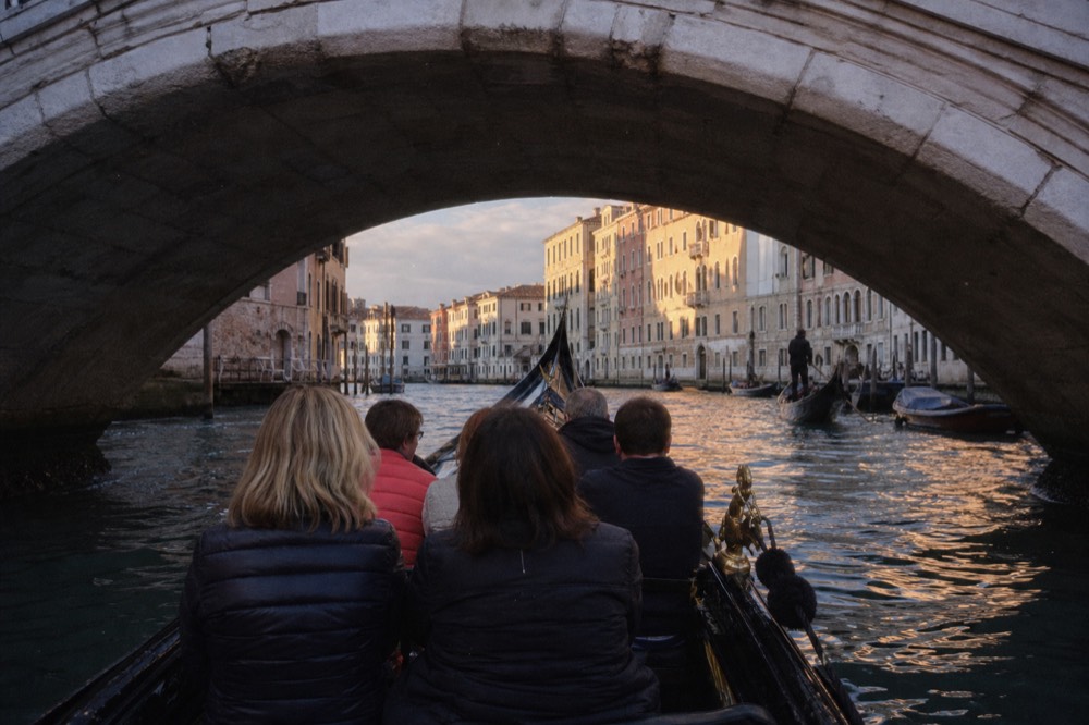 A full bench of seated passengers photographed from the stern of a gondola, backs turned, gliding under a low stone bridge in soft afternoon light