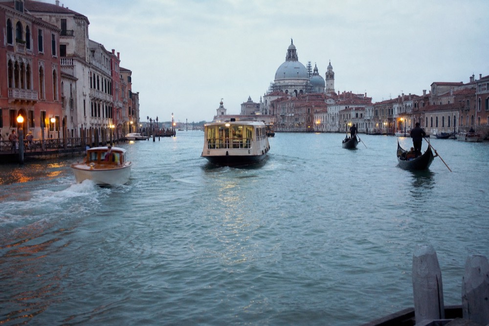 A wide stretch of open water in Venice at midday busy with a passenger ferry, a small motor launch and two gondolas riding the wake