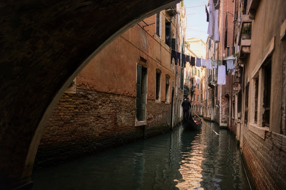 A single gondola alone on a narrow shadowed canal between tall ochre houses, laundry lines strung overhead, no other boats in sight