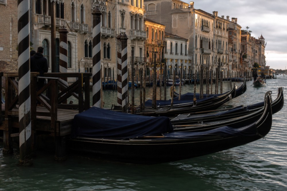 A row of moored black gondolas rocking together at a wooden landing stage, striped mooring poles leaning into green water under overcast sky