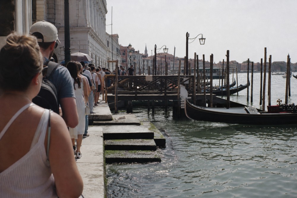 A queue of waiting travellers standing along a stone quayside in bright summer sun beside an empty gondola berth, water lapping at the steps