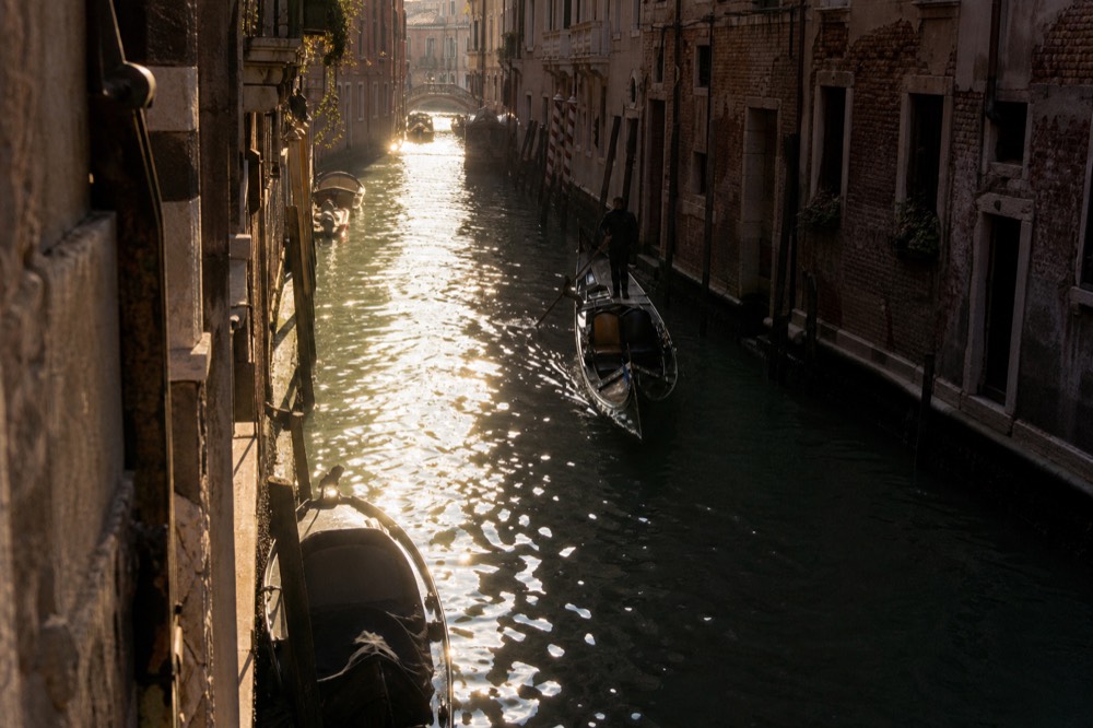 Low golden light striking one side of a narrow canal while the opposite wall stays in shadow, an empty gondola drifting through the bright strip