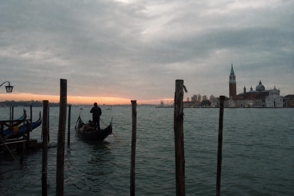 The last band of orange light on the horizon over open lagoon water with mooring poles in the foreground and a distant bell tower