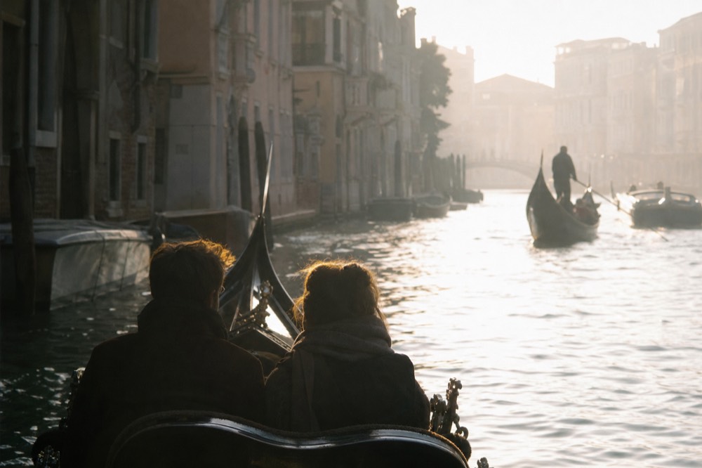 Two seated figures backlit in a gondola against a glowing water surface, their outlines sharp and the canal beyond dissolving into warm haze